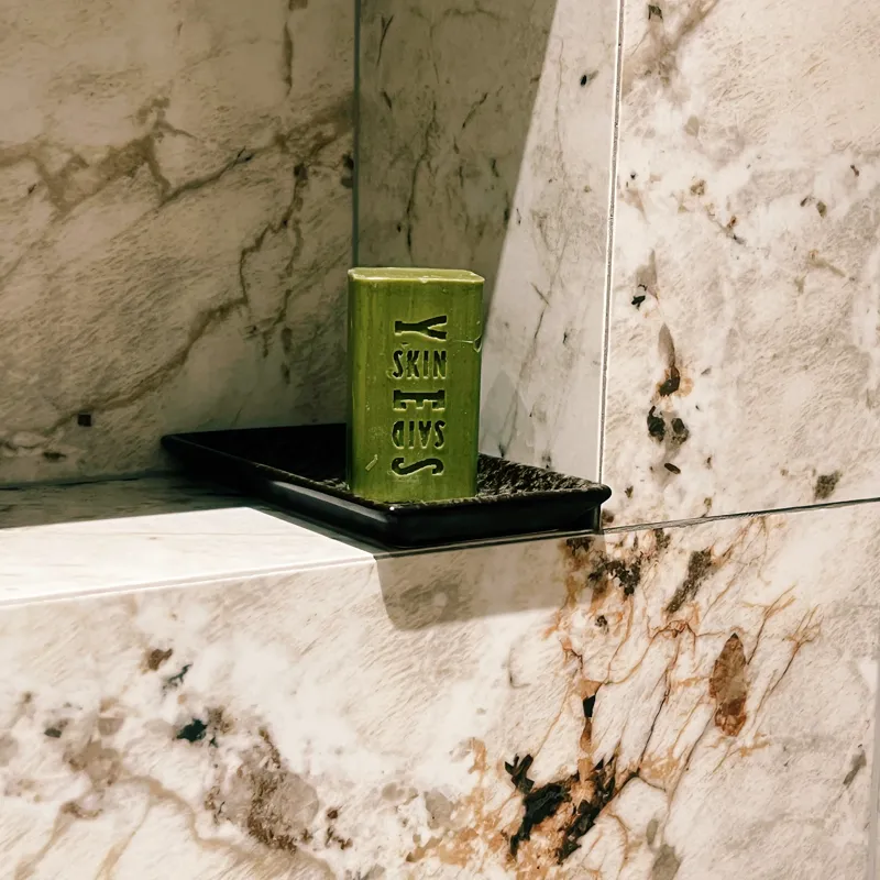 A Skin Said Yes olive oil soap bar standing on a raised dish on a shower shelf, out of the spray