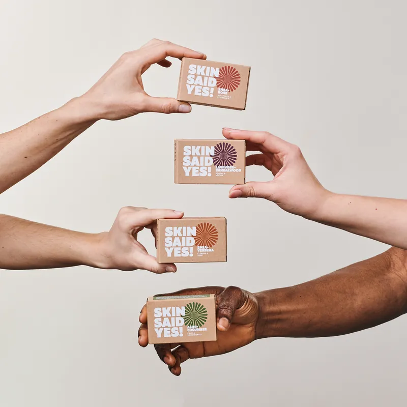 Four hands each holding a Skin Said Yes carton, one per scent